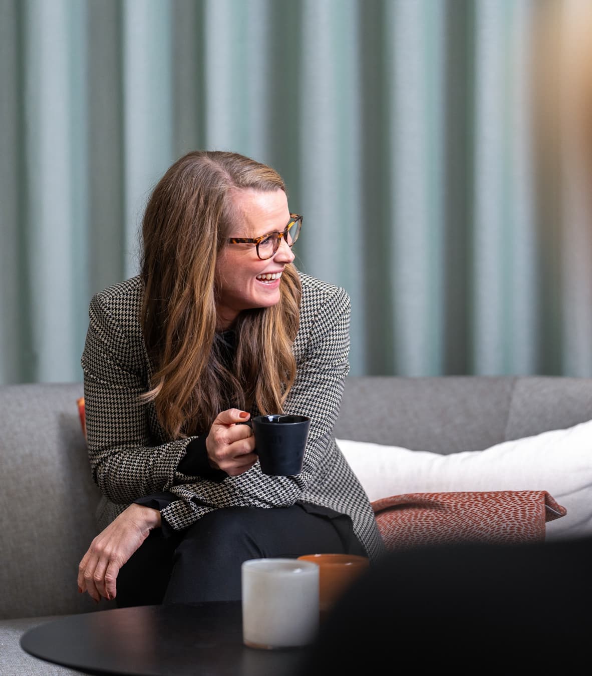 Person som sitter i soffa och skrattar med kaffekopp i handen.